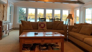 A cozy living room with a wooden ceiling, large windows, a sofa, a coffee table, and a lamp. - Maine Lake Cottage on Beech Hill Pond near Bar Harbor and Acadia National Park (Otis)