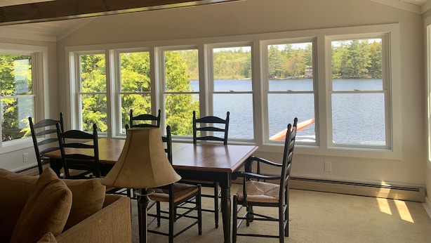 A dining area with a table and chairs, large windows with a view of a lake and trees, and a ceiling with exposed beams. - Maine Lake Cottage on Beech Hill Pond near Bar Harbor and Acadia National Park (Otis)