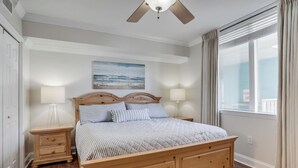 Une chambre à coucher avec un lit en bois, deux tables de chevet, un ventilateur de plafond, une fenêtre avec des rideaux et un tableau accroché au mur.