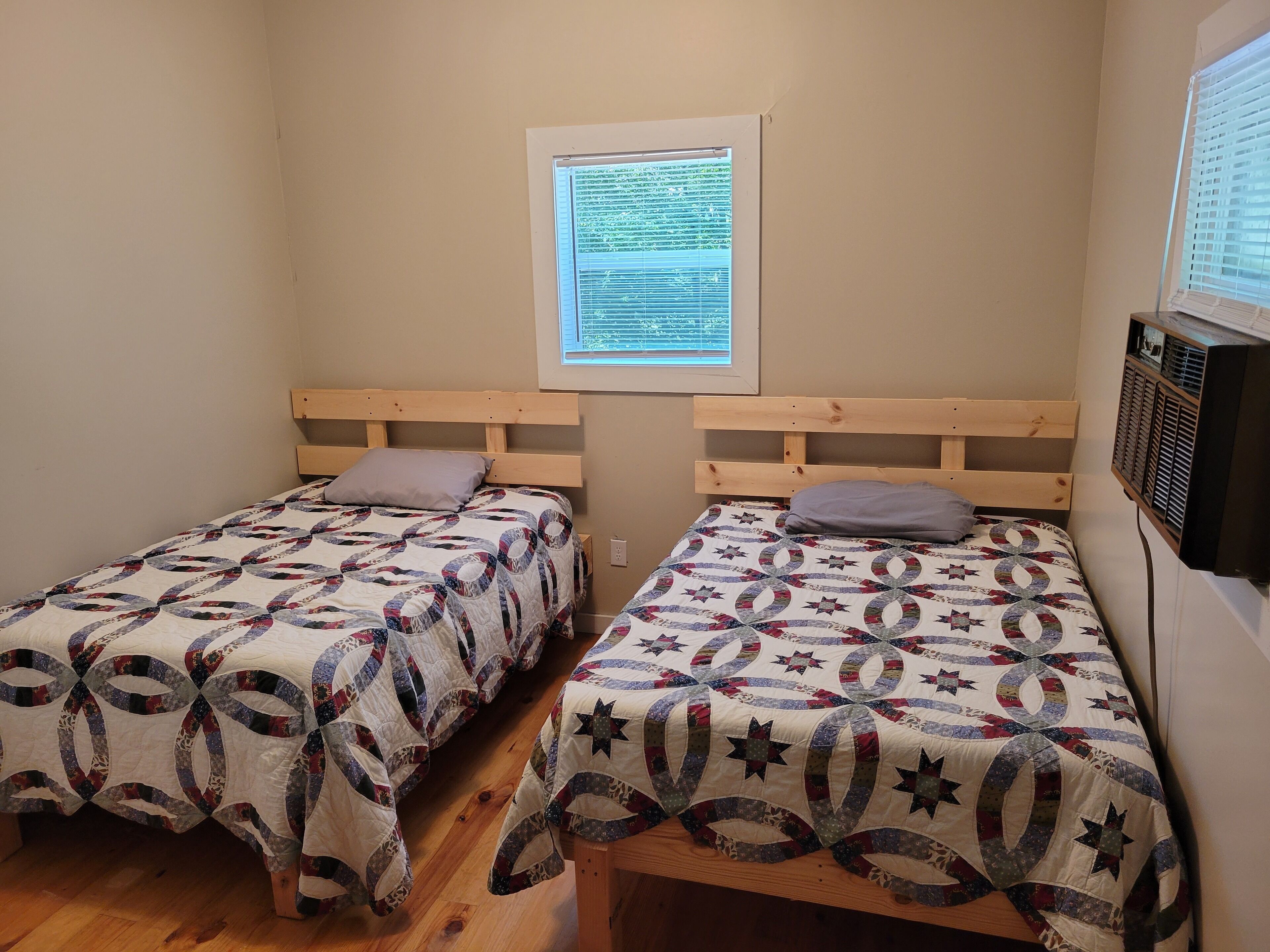 A room with two single beds, each with a wooden headboard and a quilted bedspread. There is a window with blinds, a wall-mounted TV, and a framed document on the wall.