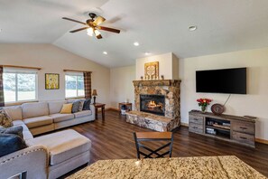 A living room with a stone fireplace, a ceiling fan, a flat-screen TV, and a large window.