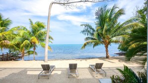 A beach with three metal lounge chairs and a white umbrella, palm trees, and a clear view of the ocean.