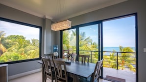 A dining area with a table and chairs, large windows with a view of the ocean and palm trees, and a water dispenser.
