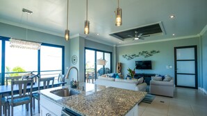 A modern kitchen with granite countertops, a dining area with wooden chairs, a living room with a sofa and TV, and a balcony with a view. - Oceanfront Sunset Gecko Condo - Ground Floor 1A (Placencia)