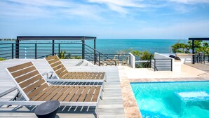 A wooden deck with lounge chairs and a pool, overlooking a clear blue sea. - Oceanfront Sunset Gecko Condo - Ground Floor 1A (Placencia)