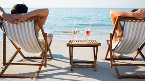 Zwei Personen sitzen auf gestreiften Liegestühlen, zum Meer zehend, mit einem kleinen Tisch dazwischen, auf dem Getränke stehen.