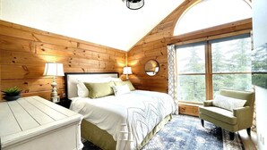 A cozy bedroom with a wooden wall, a large bed, a chair, and a window with a view of trees. - Ski In/Out, Watch Slope frm Hot Tub, 6BR, 3 Kings, PS5, Tesla, Rec Rm-Loggers 12 (Snowshoe)