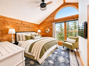 A bedroom with a bed, two bedside lamps, a green armchair, a large window with a view of trees, and a ceiling fan.
