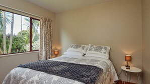A bedroom with a bed, a nightstand, a lamp, and a window with curtains. - Spacious 1 bedroom Cottage (Kaikoura)