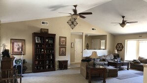 A spacious living room with a high ceiling, two ceiling fans, a large bookshelf, a sofa, a coffee table, and a dining area with chairs.