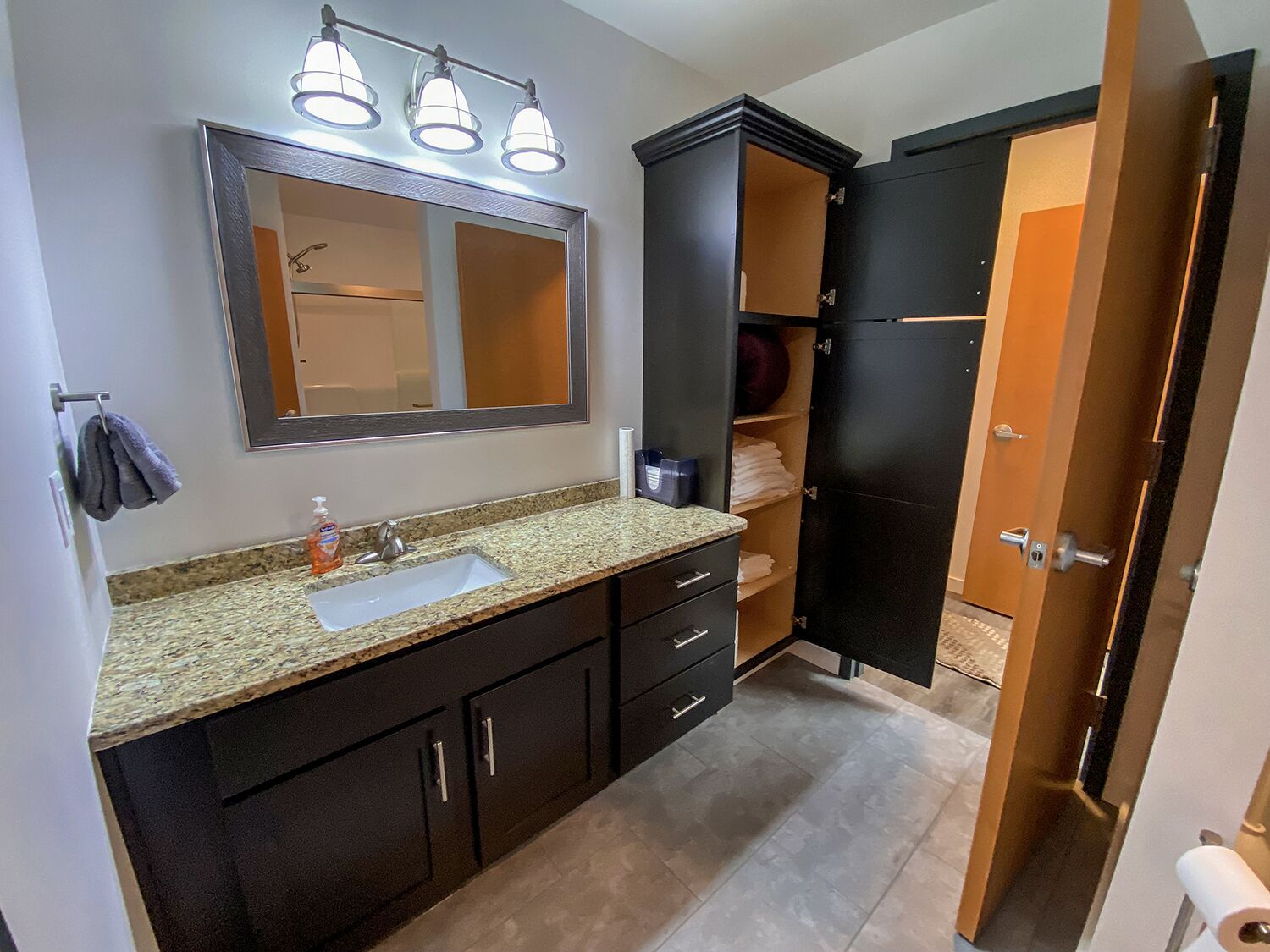 A bathroom with a granite countertop sink, a large mirror, and a dark cabinet with open doors revealing towels and toiletries.