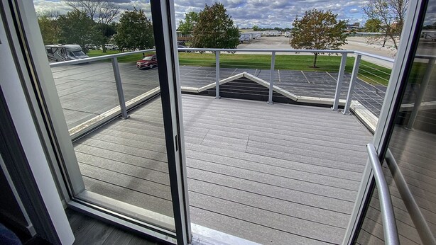 Uitzicht vanaf een balkon met een houten terras, een metalen hekwerk en een parkeerplaats met voertuigen en bomen op de achtergrond.