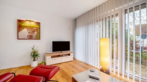 A modern living room with a flat-screen TV, red leather chairs, a wooden cabinet, a potted plant, and a large window with blinds. - Holiday Apartment at the "Gustavsburg" Castle with Wi-Fi & Terrace (Homburg)