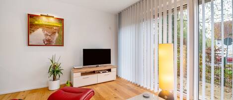 Una sala de estar moderna con un televisor de pantalla plana, sillones de cuero rojo, un mueble de madera, una planta en maceta y un ventanal con persianas.