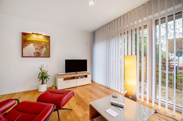 Una sala de estar moderna con un televisor de pantalla plana, sillones de cuero rojo, un mueble de madera, una planta en maceta y un ventanal con persianas.