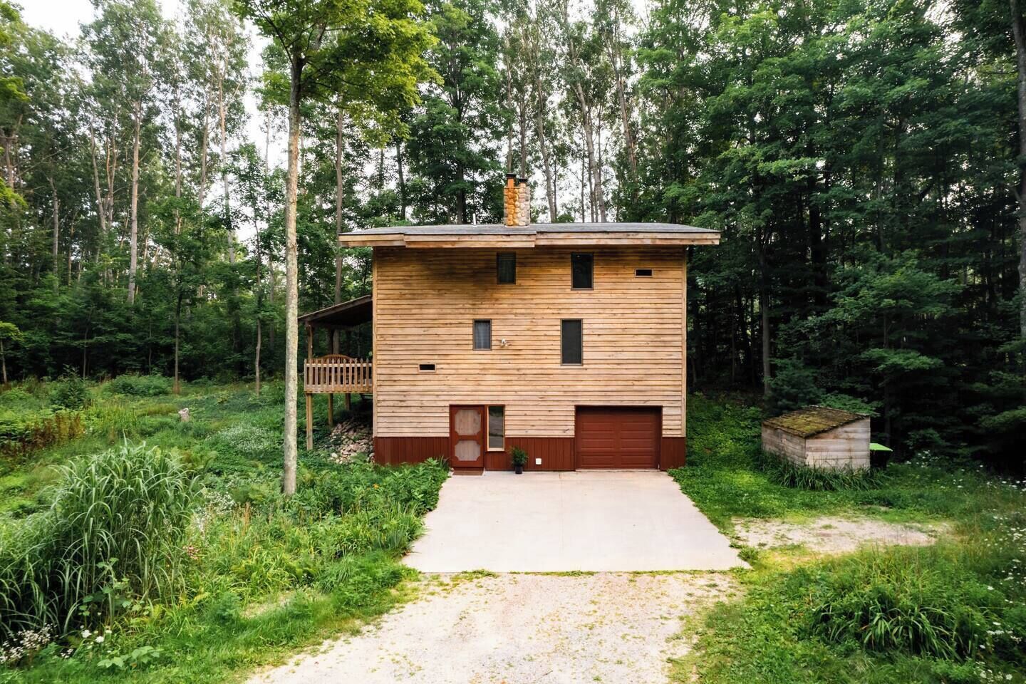 Una cabaña de madera de dos pisos con garaje, rodeada de un bosque denso.