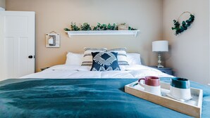 Una cama bien hecha con un edredón verde azulado, almohadas blancas y una bandeja con dos tazas.
