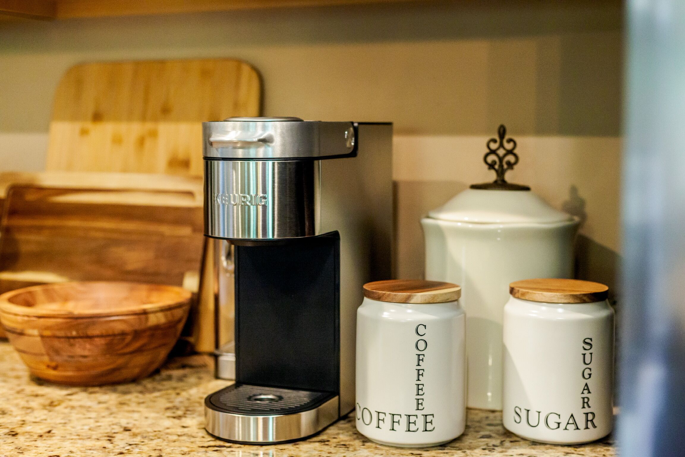 A coffee maker, two containers labeled "Coffee" and "Sugar," and a wooden bowl on a kitchen counter.