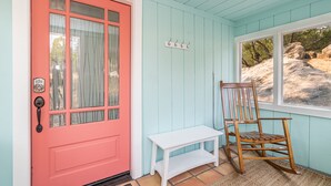 Una puerta rosa con un ventanal, una mecedora de madera y un banco blanco pequeño.