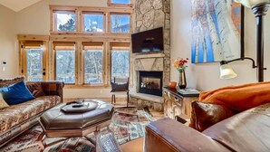 A cozy living room with a stone fireplace, large windows, a flat-screen TV, and a comfortable seating area. - Beautiful 3 bedroom with private hot tub, walkable to town and trails (Frisco)