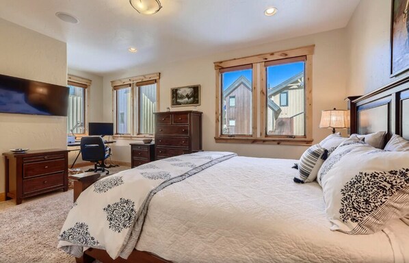 A bedroom with a large bed, a desk, and a chair. There are windows with a view of buildings outside.