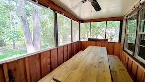 Een overdekt terras met een houten tafel en banken, grote ramen en een plafondventilator.