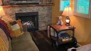 A cozy living room with a stone fireplace, a television mounted on the wall, and a window with blinds.