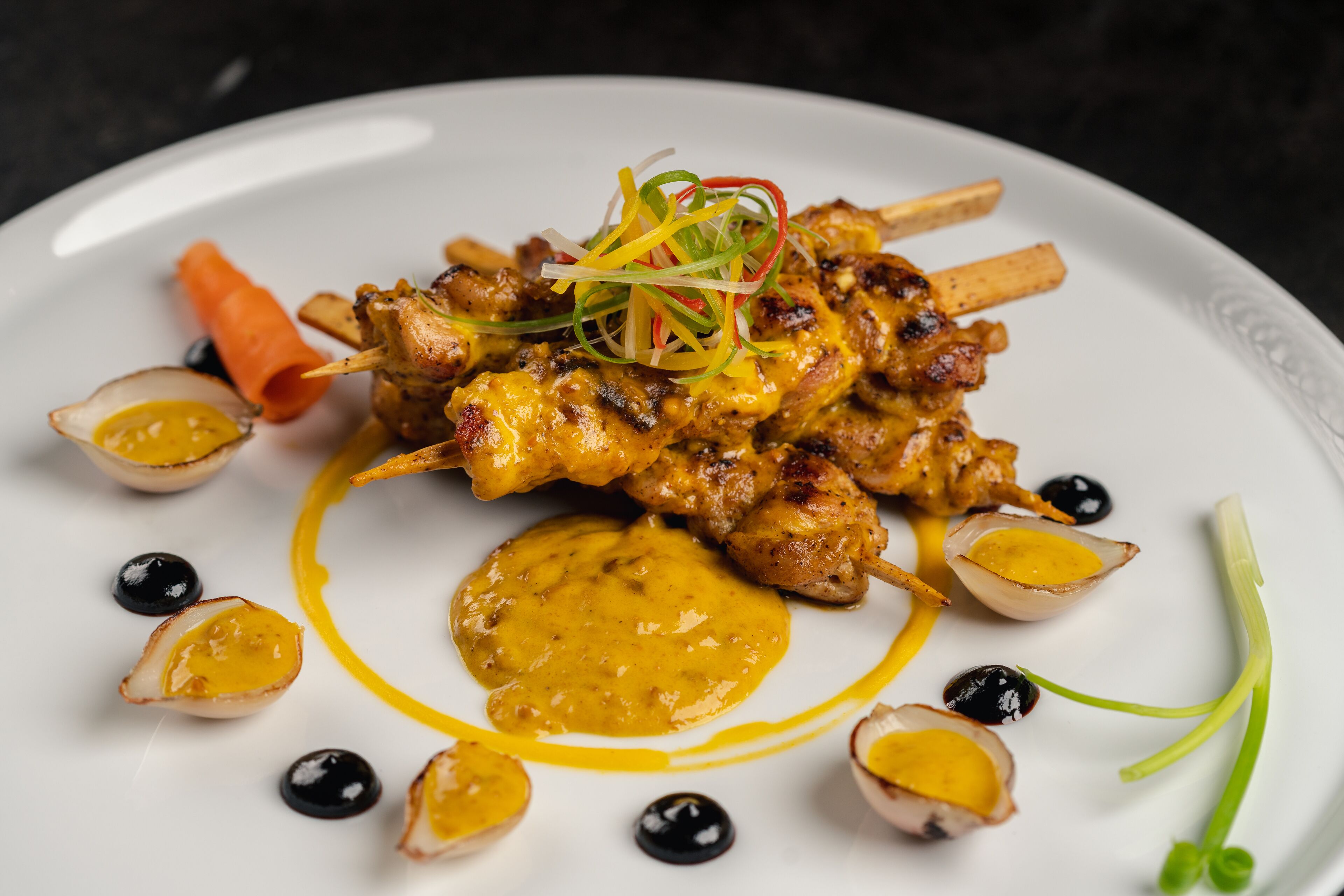 A plate of grilled chicken skewers with a yellow sauce, garnished with black olives and carrot slices.