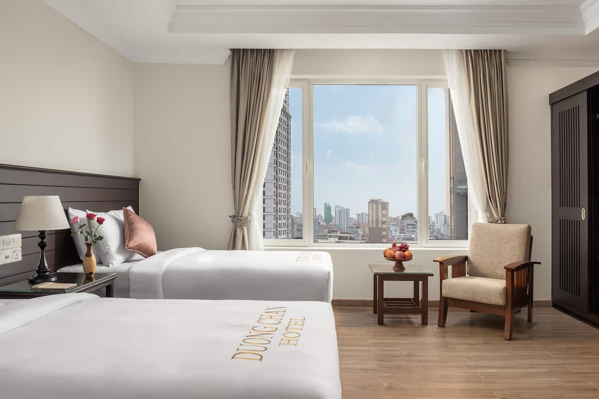 A hotel room with two beds, a nightstand with a lamp, a small table with fruit, a chair, and a view of the city skyline through large windows.