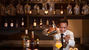 A bartender pouring a drink into a glass at a bar with various bottles and glasses in the background. - Duong Chan Hotel (Phnom Penh)