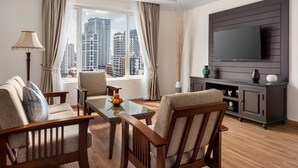 A modern living room with a glass coffee table, wooden chairs, a flat-screen TV mounted on a dark wooden panel, and a view of high-rise buildings through large windows. - Duong Chan Hotel (Phnom Penh)