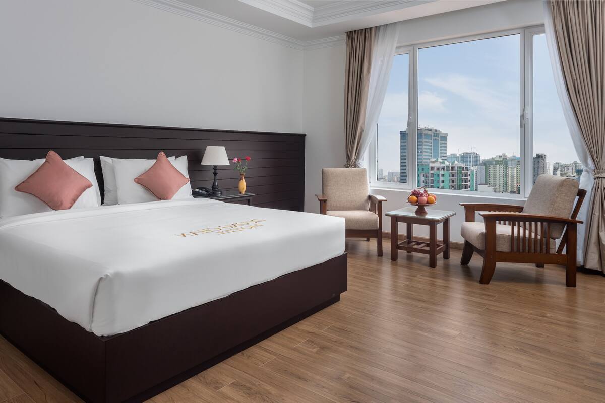 A hotel room with a large bed, two chairs, a small table, and a view of the city through large windows.