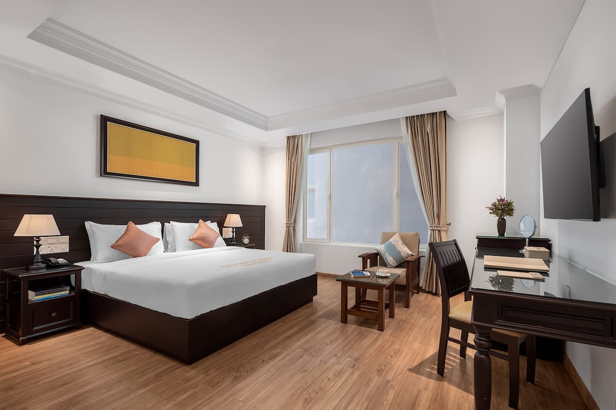 A hotel room with a large bed, bedside tables, a desk, a chair, a TV, and a window with curtains.