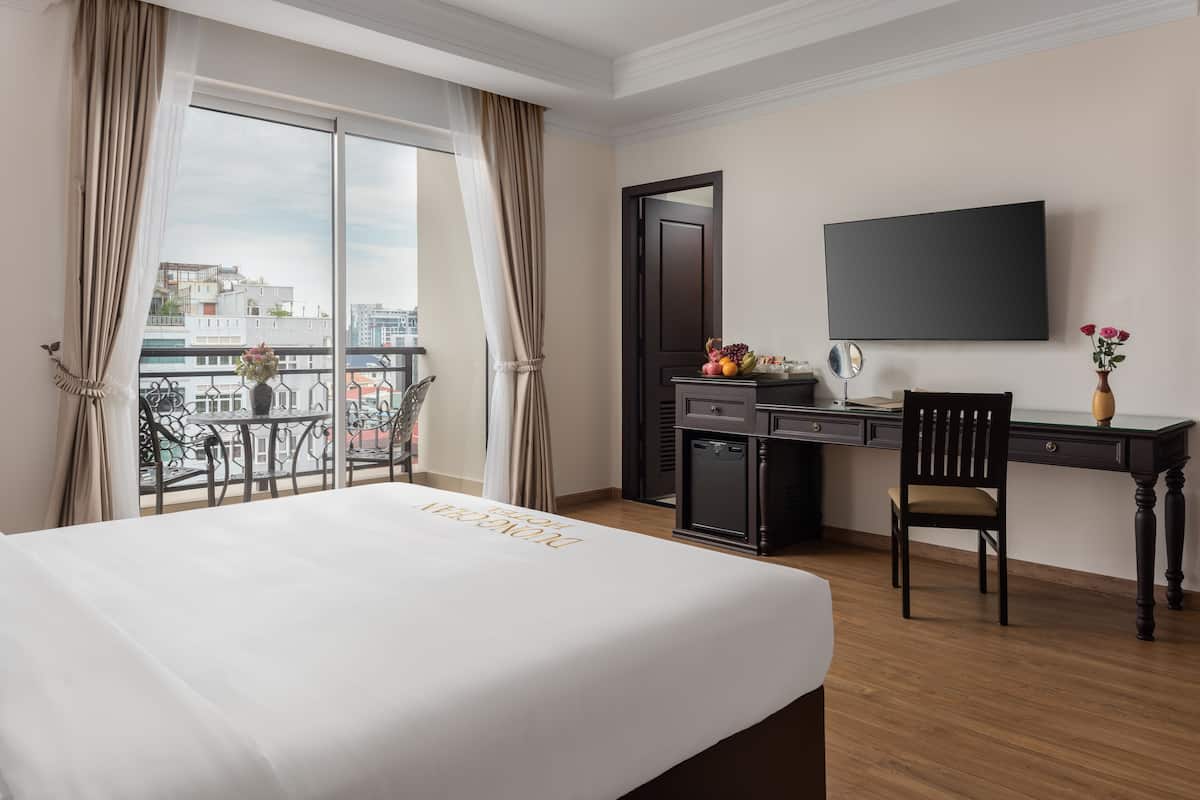 A hotel room with a bed, a desk, a chair, a TV, and a balcony with a view of buildings.