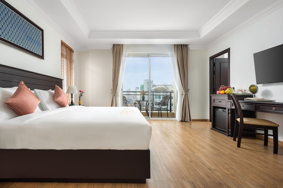 A hotel room with a large bed, a desk with a chair, a balcony with a view of the city, and a TV on the wall.
