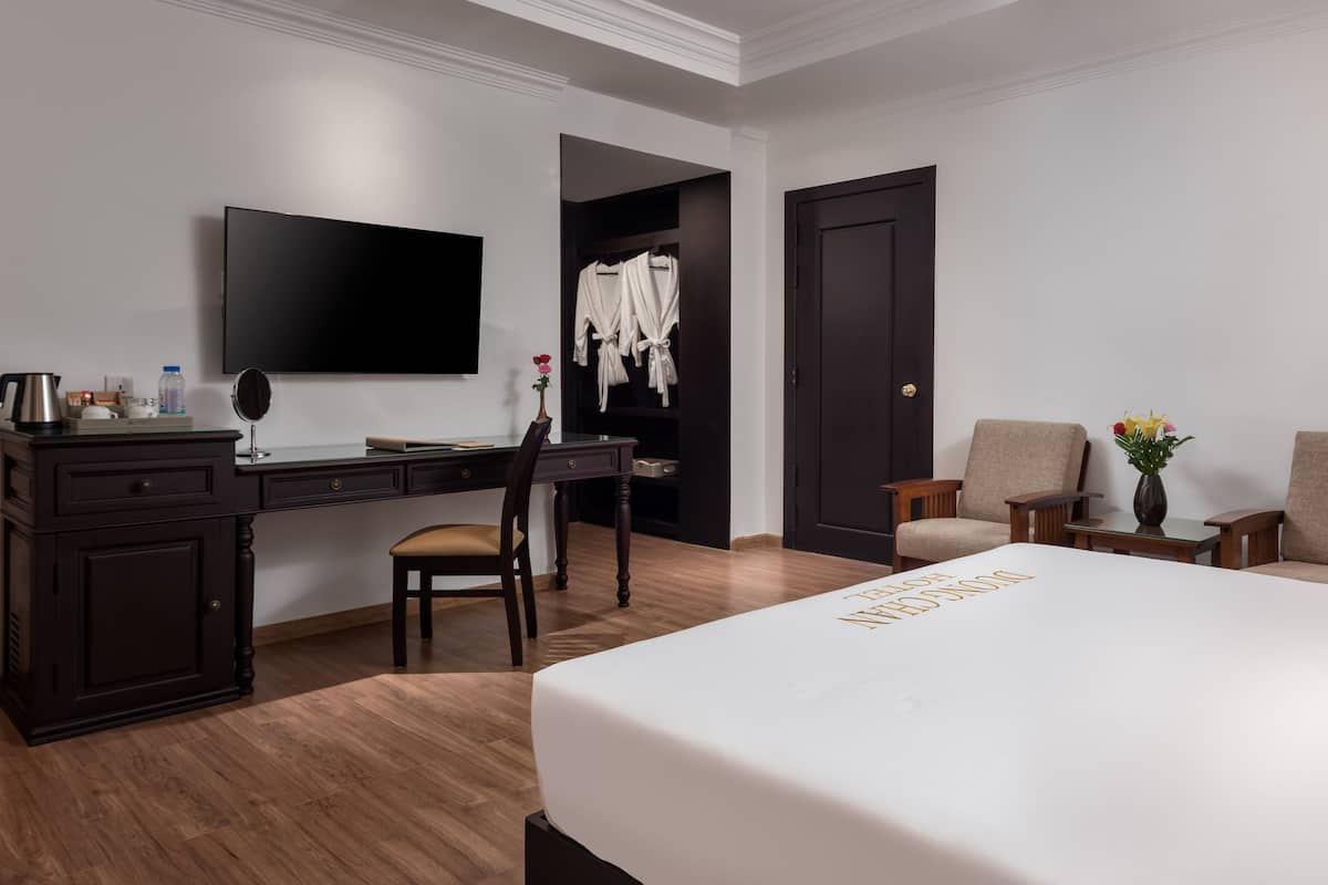 A hotel room with a large bed, a desk with a chair, a television, a wardrobe, and a seating area with two chairs.