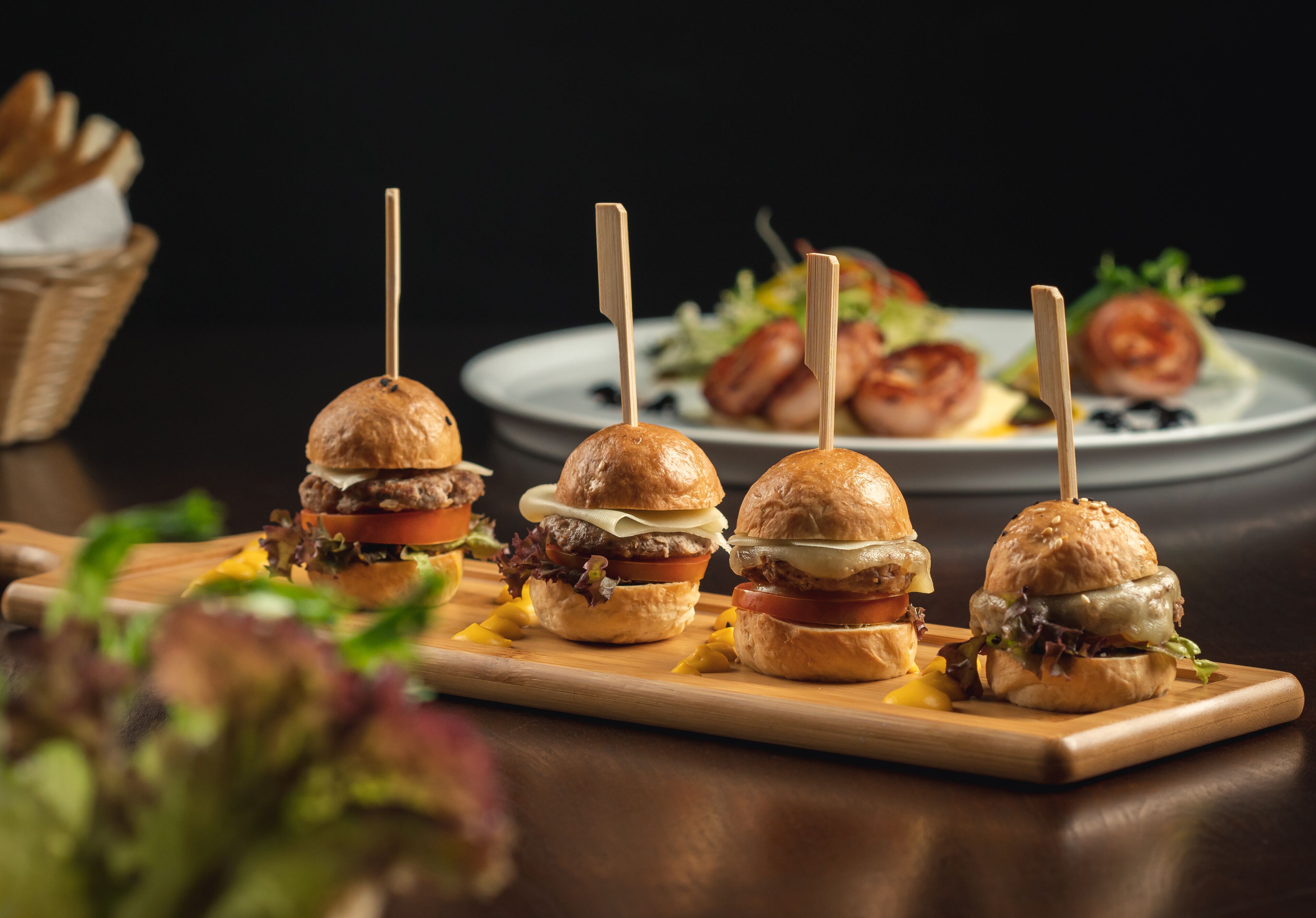 Een houten plank met vier burgers op spiesjes, een broodmand en een bord eten op de achtergrond.