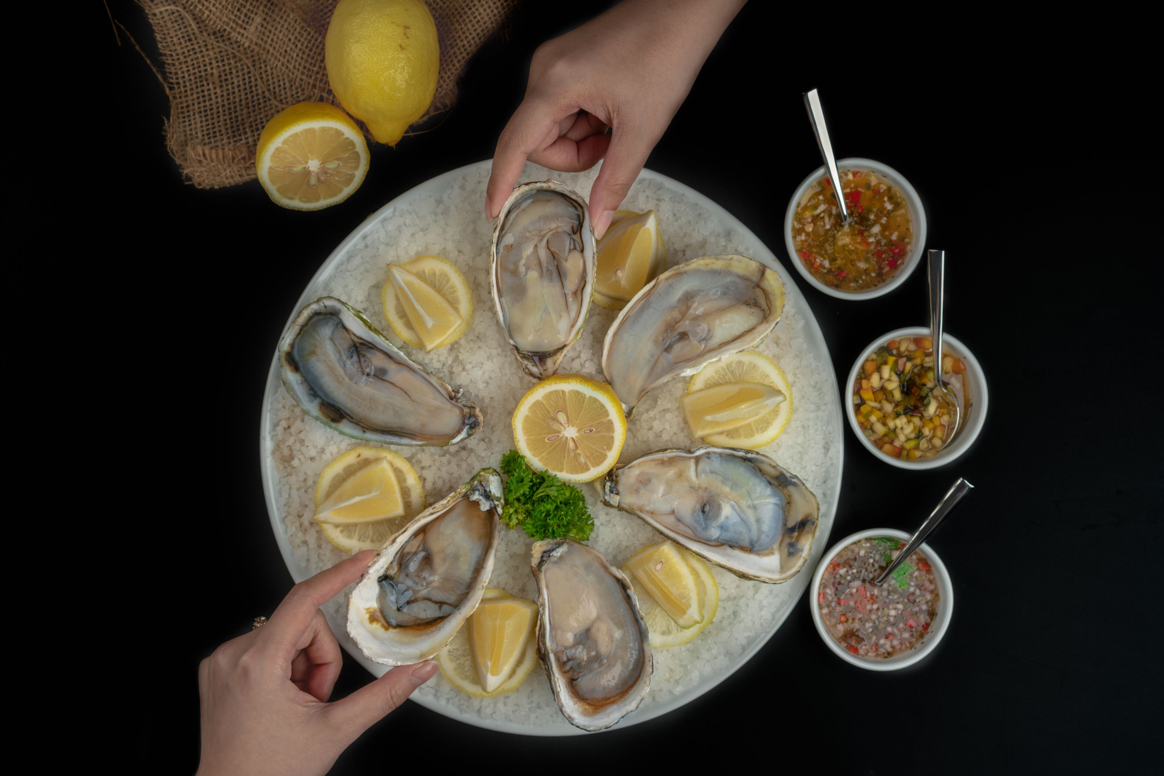 Un plato de ostras con rodajas de limón y perejil, acompañado por dos recipientes pequeños de salsas.