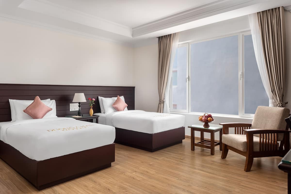A hotel room with two beds, a wooden floor, a window with curtains, and a chair.