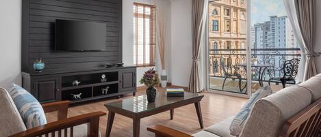 A living room with a TV mounted on a dark wall, a glass coffee table, wooden furniture, and a balcony with a view of buildings.