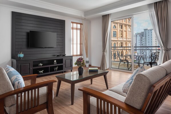 A living room with a TV mounted on a dark wall, a glass coffee table, wooden furniture, and a balcony with a view of buildings.