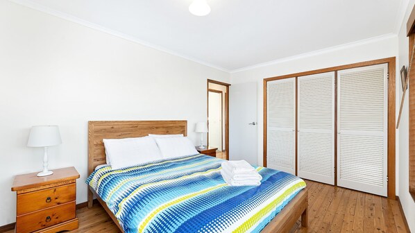 A bedroom with a wooden bed, white pillows, a striped bedspread, a wooden nightstand with a lamp, and a wooden wardrobe with louvered doors. - 27 Harrison Street, Dalmeny (Dalmeny)
