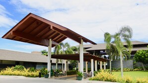 A covered outdoor area with a wooden roof, surrounded by greenery and a building in the background. - Cocoons Club Laiya (San Juan)