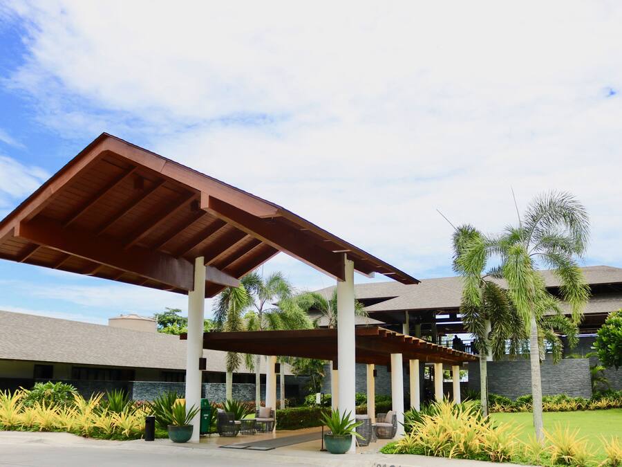 A covered outdoor area with a wooden roof, surrounded by greenery and a building in the background.