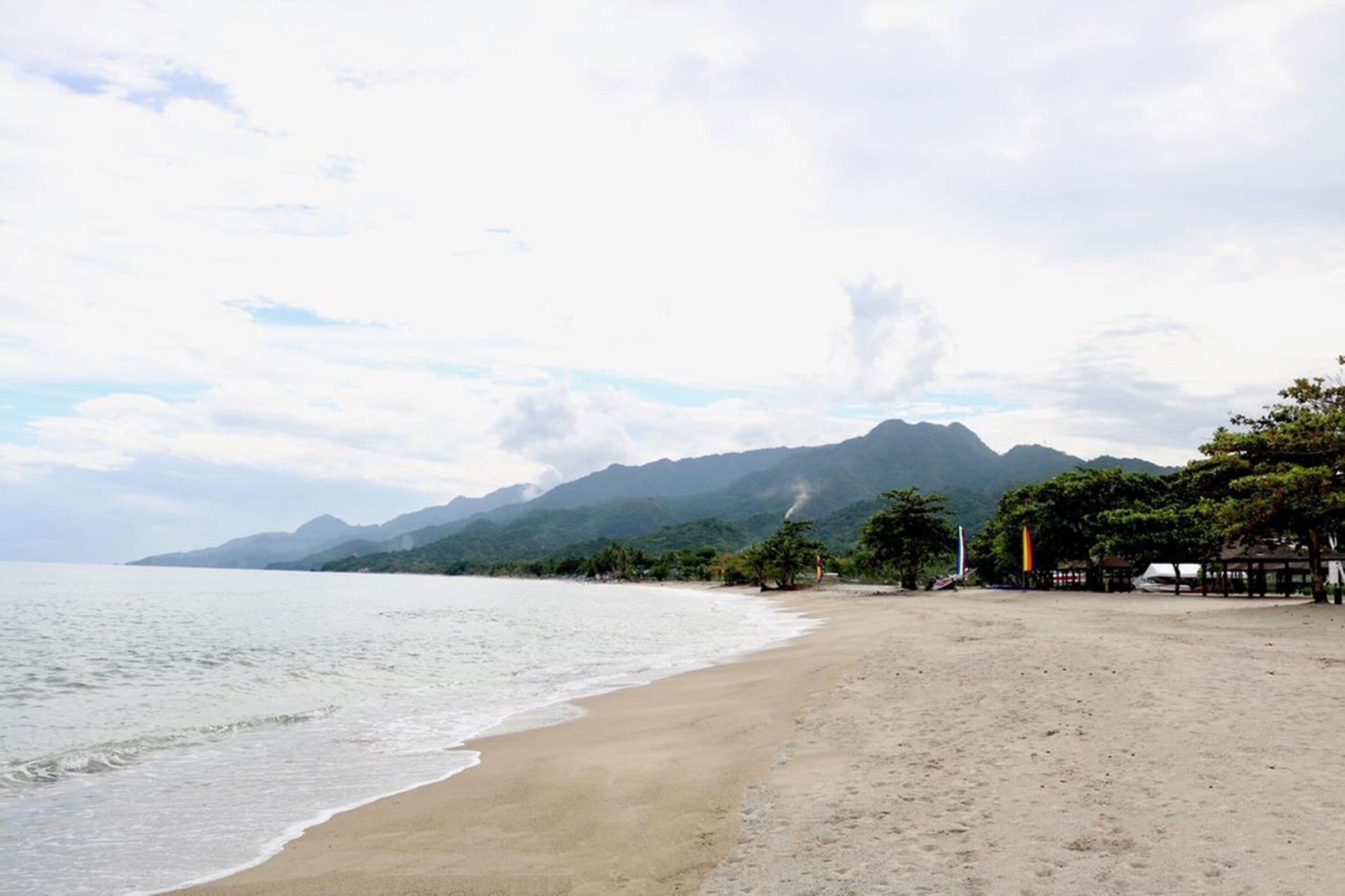 Una playa con una montaña al fondo, una orilla arenosa y algunos árboles.