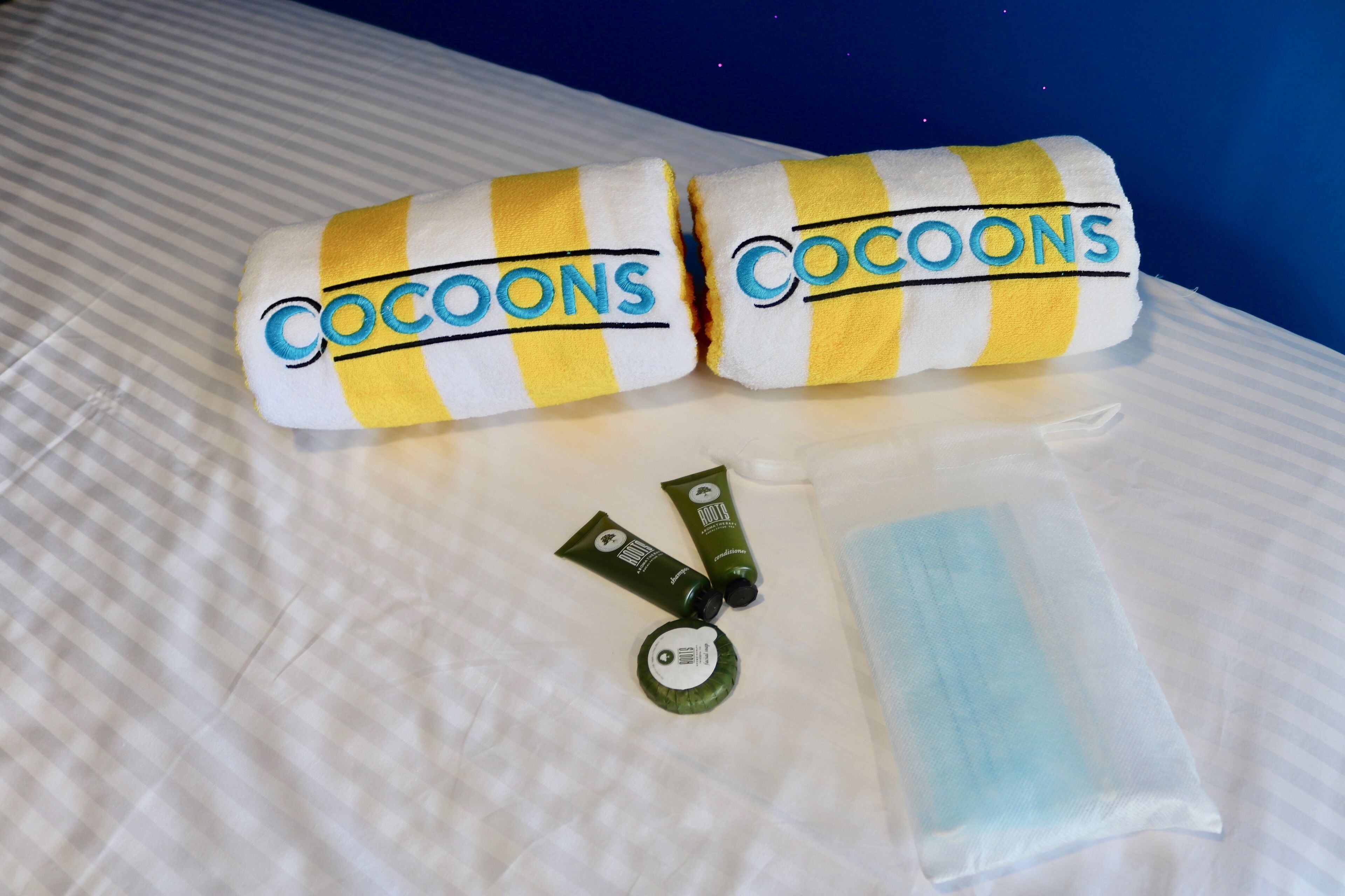 Dos toallas enrolladas con la marca 'COCOONS', un tubo verde y una mascarilla azul sobre una cama.