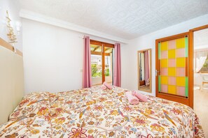 Une chambre à coucher avec une couvre-lit à motifs floraux, une porte en bois ornée d’un panneau de verre coloré, et une vue sur l’extérieur.