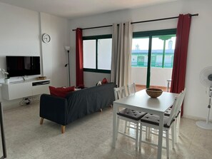 Un salon moderne doté d’un téléviseur à écran plat, d’un canapé, d’une table à manger avec des chaises, d’un ventilateur et d’une grande fenêtre avec des rideaux.