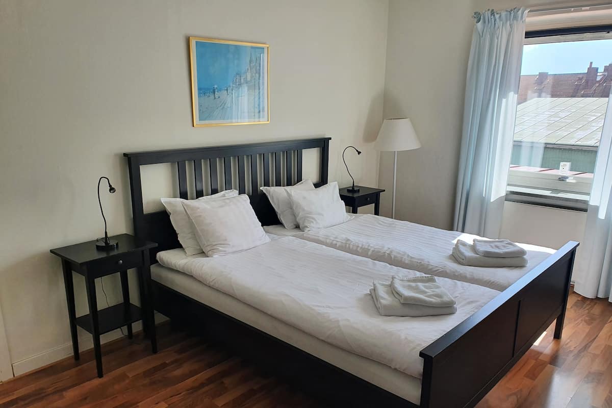 Un lit double avec du linge de lit blanc, deux tables de chevet, une commode, une lampe, un tableau au mur et une fenêtre avec des rideaux.
