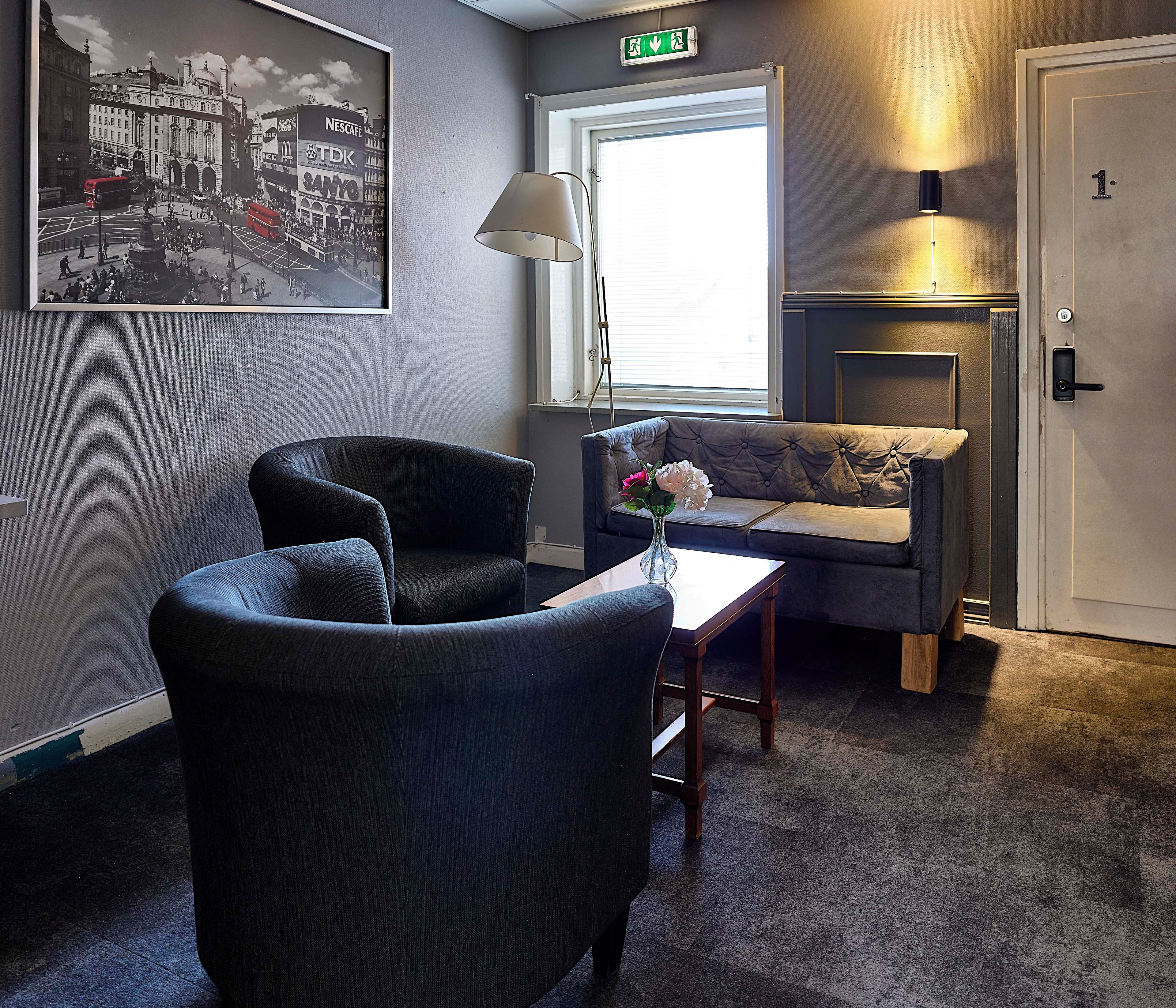 Ett hotellrum med en sittgrupp bestående av två fåtöljer och en soffa, ett litet bord med en vas, en öppen spis och en inramad tavla som föreställer en stadsbild på väggen.